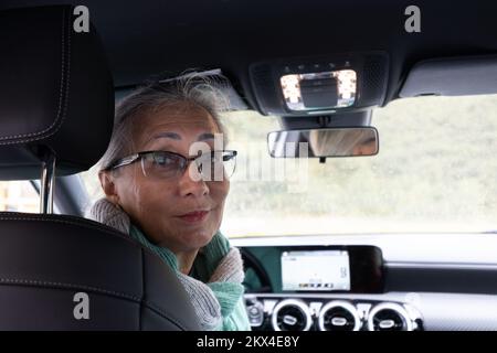 Eine grauhaarige Dame sitzt auf dem Fahrersitz eines Autos und schaut sich die Passagiere an. Stockfoto