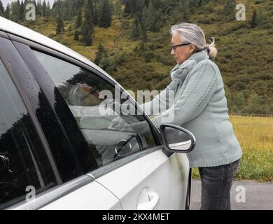 Eine Erwachsene grauhaarige Frau in einem warmen Pullover und einer Brille stellt die Scheibenwischer eines Autos ein Stockfoto
