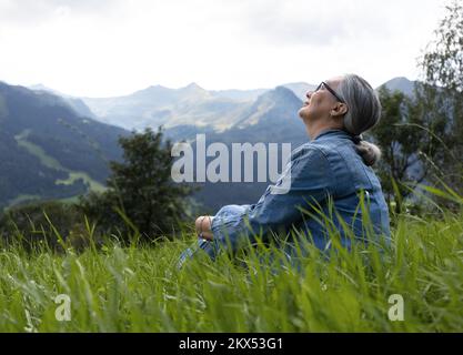 Eine ältere Frau im Jeansanzug sitzt auf einer alpinen Wiese und schaut in den Himmel Stockfoto