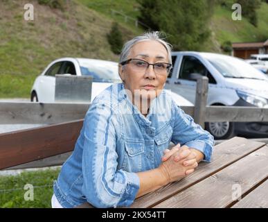 Eine grauhaarige Frau im Jeansanzug sitzt auf einer Bank in der Nähe eines Parkplatzes. Stockfoto
