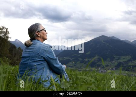 Eine ältere Frau im Jeansanzug sitzt auf einer alpinen Wiese und blickt auf die Berge. Stockfoto