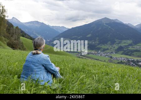 Eine grauhaarige Frau in einem Jeansanzug sitzt auf einem grasbedeckten Hang und blickt auf die Berge. Stockfoto