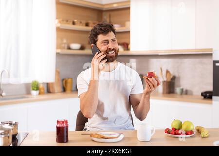 Ein erwachsener, weißer Kerl mit Bart im weißen T-Shirt isst Sandwich, telefoniert in einer modernen Küche Stockfoto