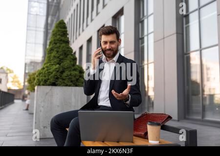 Lächelnder junger europäischer Manager mit Bart im Anzug, einer Tasse Kaffee und Telefonaten mit dem Laptop Stockfoto