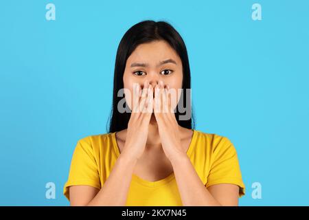 Das Porträt einer schockierten Asiatin im gelben T-Shirt bedeckt ihren Mund mit Händen und schaut in die Kamera Stockfoto