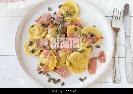 Ravioli mit Pilzfüllung, gekocht mit italienischem Schinken, Butter und gerösteten Kürbiskernen. Serviert auf einem weißen Teller, isoliert auf hellem Holzhintergrund Stockfoto