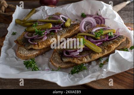 Gebratener Hering mit roten Zwiebeln, Gurken auf buttertem und geröstetem Roggenbrot auf einem Holztisch Stockfoto