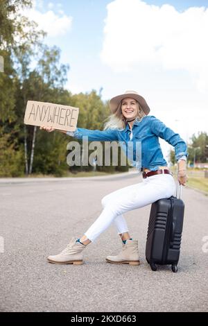 Fröhliche Frau hört auf, an einem Auto vorbeizuschauen, sitzt auf einem Koffer mit einem ausgestreckten Pappposter auf einer leeren Autobahn. Lady mit Hut, entfliehen Sie der Stadt, um überall hinzugehen Stockfoto