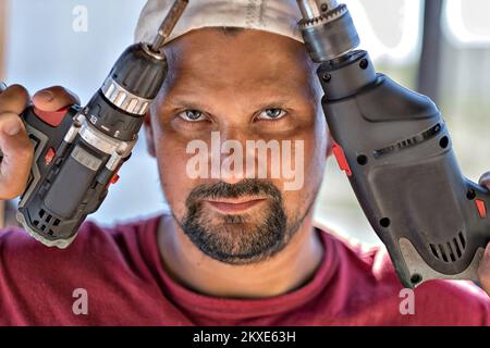 Ein männlicher Schmalzbauer bei der Arbeit. Nahaufnahme eines Arbeiters mit Bart und Mütze. Das Gesicht hat Werkzeuge, einen Schraubendreher und einen Bohrer. Hochwertiges Foto. Stockfoto