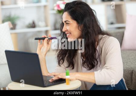 Reife Frau, die am Laptop arbeitet und eine elektronische Zigarette raucht Stockfoto