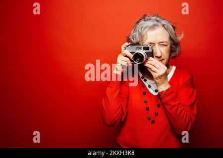 Ältere Frau in roter Kleidung, fotografiert mit einer alten Filmkamera, auf rotem Hintergrund Stockfoto