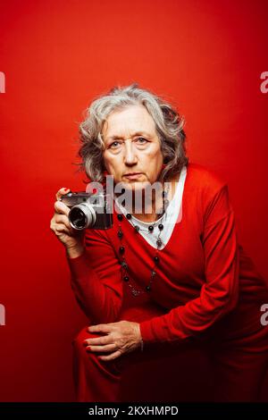 Ältere Frau in roter Kleidung, fotografiert mit einer alten Filmkamera, auf rotem Hintergrund Stockfoto