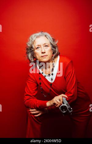 Ältere Frau in roter Kleidung, fotografiert mit einer alten Filmkamera, auf rotem Hintergrund Stockfoto