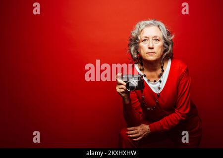 Ältere Frau in roter Kleidung, fotografiert mit einer alten Filmkamera, auf rotem Hintergrund Stockfoto