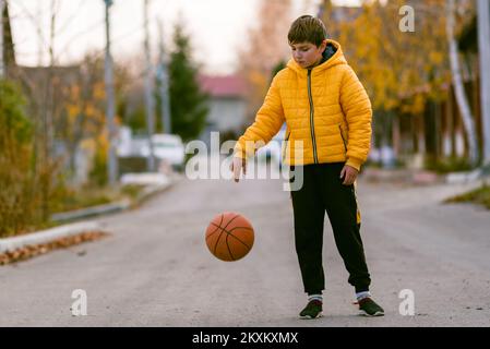 Junger Junge, der in den Händen hält Basketballsport Junge, kleiner Kaukasier, der den orangefarbenen Ball spielt, dribbelnde, gelb gepolsterte Jacke o Stockfoto