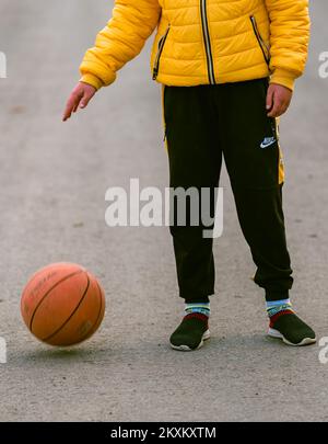 Junger Junge, der in den Händen hält Basketballsport Junge, kleiner Kaukasier, der den orangefarbenen Ball spielt, dribbelnde, gelb gepolsterte Jacke o Stockfoto