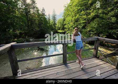 Die Rückseite des Mädchens steht auf der Holzbrücke des smaragdgrünen Wasserflusses Sava Bohinjka in den Julischen Alpen, Ukanc, Slowenien. Stockfoto