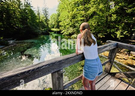 Die Rückseite des Mädchens steht auf der Holzbrücke des smaragdgrünen Wasserflusses Sava Bohinjka in den Julischen Alpen, Ukanc, Slowenien. Stockfoto