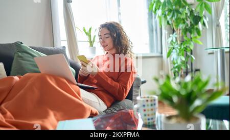Entspannte junge Frau, die auf dem Sofa sitzt und ein Telefon in einem gemütlichen Wohnzimmer hält. Stockfoto
