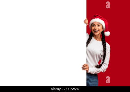 Wunderschöne Araberin In Santa Hat, Die Neben Dem Blank White Advertisement Board Steht Stockfoto