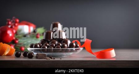 Stapel von Schokoladenbonbons auf Glasplatte mit roter Schleife und Weihnachtsdekoration auf dunklem, isoliertem Hintergrund. Vorderansicht. Stockfoto