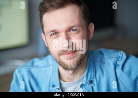 Attraktiver Mann in Jeansshirt, positive Emotionen mit Computermonitor im Hintergrund. Porträt eines fröhlichen bärtigen männlichen Software-Ingenieurs, der am Arbeitsplatz im Büro sitzt und in die Kamera schaut Stockfoto