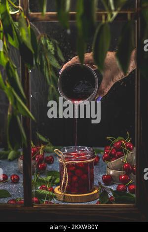 Hausgemachte Konfitüre aus roter Kirschkonfitüre in Dosen mit Chili im Glasglas, die auf einem dunklen Holztisch hinter dem Fenster steht Stockfoto