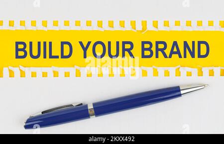 Wirtschaft und Finanzen. Auf dem Tisch befinden sich zwei Blätter aus einem Notizbuch und ein Stift auf gelbem Hintergrund mit der Aufschrift „BAUEN SIE IHRE MARKE“ Stockfoto