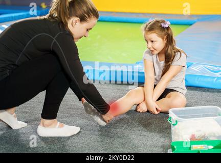 Ein kleines Mädchen hat Schmerzen im Knöchel und bekommt Hilfe, nachdem es auf Trampolin gesprungen ist, roter Fleck zeigt Schmerzen Stockfoto