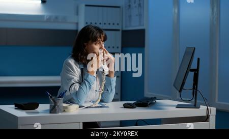 Arzt Arzt am Festnetztelefon mit entferntem Patienten diskutieren medizinische Verschreibung erklären medizinische Behandlung. Fachfrau, die über Stunden in der Krankenhauspraxis arbeitet Stockfoto