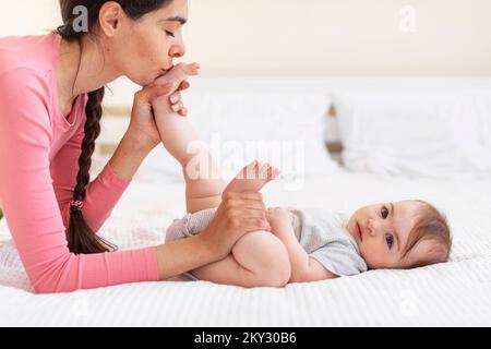 Pflegende junge Mutter küsst die Füße des Babys, während sie zu Hause im Bett Gymnastik macht, Seitenansicht, Kopierbereich Stockfoto