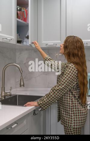 Süße junge Hausfrau öffnet die Tür zum Küchenschrank Stockfoto