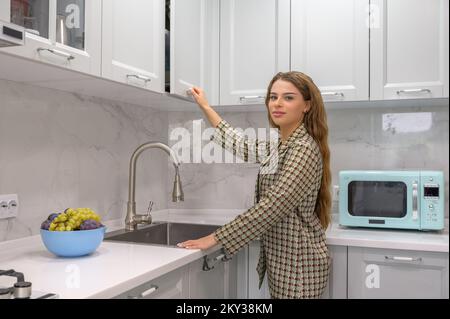 Süße junge Hausfrau öffnet die Tür zum Küchenschrank Stockfoto