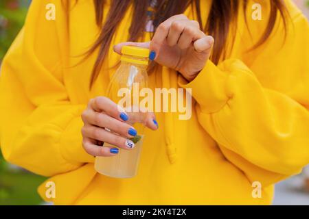 Eine junge, nicht erkennbare Frau öffnet an einem Sommertag draußen eine Plastikflasche Zitronensaft und trägt einen gelben Hoodie vor einem bunten Gebäude Stockfoto