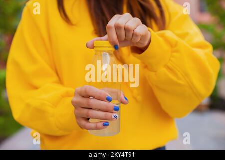 Eine junge, nicht erkennbare Frau öffnet an einem Sommertag draußen eine Plastikflasche Zitronensaft und trägt einen gelben Hoodie vor einem bunten Gebäude Stockfoto