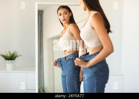 Schockierte junge Frau, die nach erfolgreicher Gewichtsabnahme zu große Jeans trägt, posiert und ihr Spiegelbild betrachtet Stockfoto