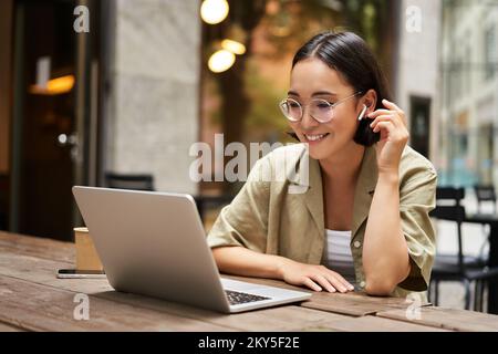 Porträt eines asiatischen Mädchens, das im Café arbeitet, einen Laptop benutzt und draußen auf der Straße sitzt. Digitales Nomaden- und Online-Lernkonzept Stockfoto