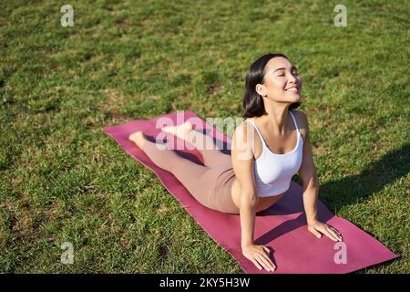 Lächelnde junge Sportlerin streckt sich im Park auf einer Gummimatte, macht Yoga-Asana-Übungen, trainiert an der frischen Luft in Fitnesskleidung Stockfoto