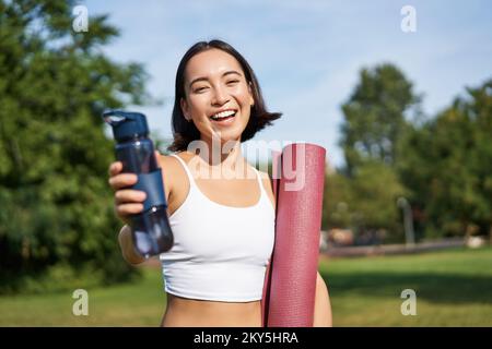 Porträt eines glücklichen asiatischen Mädchens, eine Fitnessfrau, die dir nach dem Training eine Flasche Wasser zum Trinken gibt, mit einer Gummimatte für Übungen im Park Stockfoto