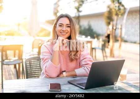 Porträt einer glücklichen, erfolgreichen, selbstbewussten Kaukasierin, Managerin, Freiberuflerin, in lässiger Kleidung, sitzt draußen in der Nähe des Büros mit einem Laptop auf dem Schreibtisch, schaut in die Kamera, lächelt freundlich Stockfoto
