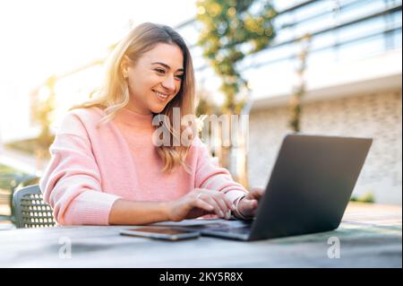 Positiv erfolgreiche junge weiße blonde Frau, Freiberufler, Studentin, sitzt draußen mit einem Laptop am Schreibtisch, arbeitet an einem Projekt, surft im Internet, beantwortet E-Mails, lächelt Stockfoto