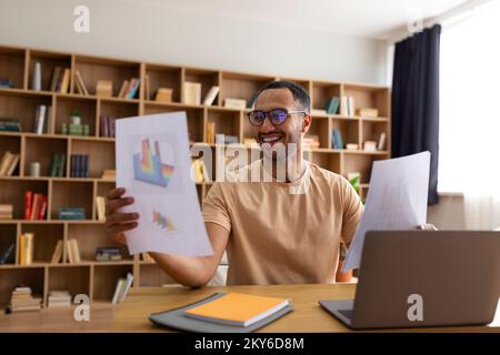 Virtuelle Besprechung. Ein glücklicher arabischer Mann, der Videoanrufe hat, Dokumente mit Diagrammen überprüft und ein Notebook benutzt Stockfoto