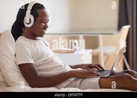 Lächelnder Schwarzer Mit Kopfhörern, Der Im Bett Auf Dem Laptop Sitzt Stockfoto