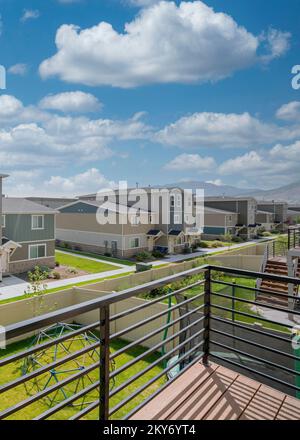 Vertikale weiße Puffelwolken Blick auf die Nachbarhäuser mit Rasen- und Betonwegen von einer Terrasse. Terrasse eines eingezäunten Hauses mit Holzboden und Stockfoto