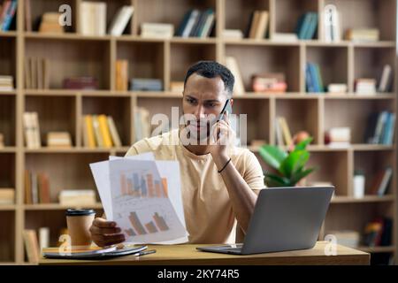 Irritierter arabischer Geschäftsmann, der von zu Hause aus arbeitet, am Schreibtisch mit einem Laptop sitzt, Dokumente in der Hand hält und telefoniert Stockfoto