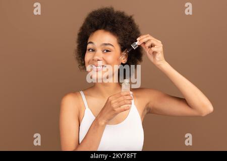 Glückliche Millennials schwarze, geschwungene Frau mit weißem Oberteil Anti-Aging-Kollagenserum auftragen, isoliert auf braunem Hintergrund Stockfoto