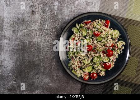 Couscous-Salat mit Gurken, Tomaten und rosafarbenen Zwiebeln auf einem runden Teller mit dunklem Hintergrund. Draufsicht, flach liegend Stockfoto