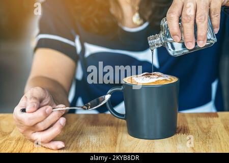 Nahaufnahme einer reifen Frau, die auf dem Kaffeetisch warmen Sirup in heißen Cappuccino gießt. Sirup in eine heiße Kaffeetasse gießen. Hinzufügen einer subtilen Süße zu favori Stockfoto