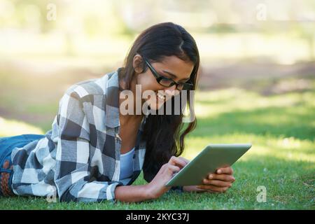 Immer in Verbindung, egal wo ich bin. Eine junge Frau, die an einem Touchpad arbeitet, während sie in einem Park liegt. Stockfoto