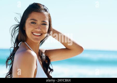 Windgepeitschte Schönheit. Eine wunderschöne junge Frau, die am Strand in der Sonne steht. Stockfoto